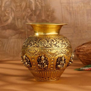 May include: A gold-colored metal vase featuring intricate embossed floral patterns and detailed relief carvings of deities around the central body. The vessel has a wide, flared rim, a rounded midsection, and a tapered base with a fluted design.
