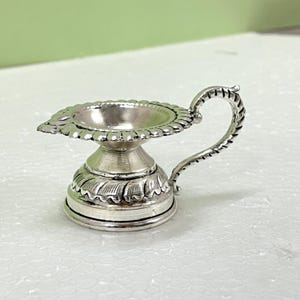 Peut inclure: Une petite lampe &agrave; huile en m&eacute;tal argent&eacute; dot&eacute;e d'un large bord festonn&eacute;, d'une base en pi&eacute;destal effil&eacute;e avec des d&eacute;tails grav&eacute;s en forme de feuilles et d'une poign&eacute;e incurv&eacute;e et textur&eacute;e s'&eacute;tendant du c&ocirc;t&eacute; du bol jusqu'&agrave; la base.
