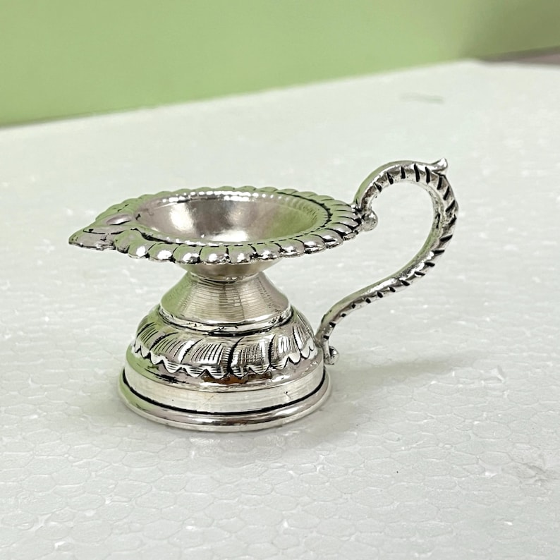 Peut inclure: Une petite lampe &agrave; huile en m&eacute;tal argent&eacute; dot&eacute;e d'un large bord festonn&eacute;, d'une base en pi&eacute;destal effil&eacute;e avec des d&eacute;tails grav&eacute;s en forme de feuilles et d'une poign&eacute;e incurv&eacute;e et textur&eacute;e s'&eacute;tendant du c&ocirc;t&eacute; du bol jusqu'&agrave; la base.