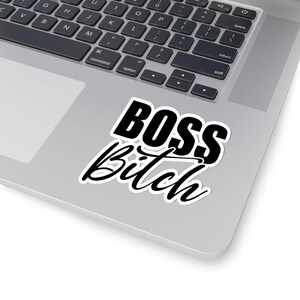 May include: A white sticker with the words "BOSS Bitch" in black, bold, and cursive font. The sticker is placed on a silver laptop, with the keyboard visible in the background.