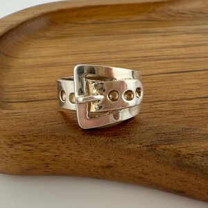 May include: A polished silver ring designed to resemble a miniature belt, featuring a rectangular buckle frame, a central prong, and a series of circular holes along the band that mimic the appearance of a functional leather belt strap.