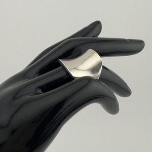 May include: A silver ring with a curved, wave-like design. The ring is smooth and polished, reflecting light. The ring is shown on a black hand, creating a contrast that highlights the jewelry's shape and shine. The ring is a piece of fashion jewellery.