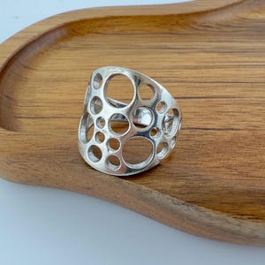 May include: A silver-toned ring featuring a wide band with a cut-out design composed of multiple circular openings of varying sizes. The metal surface is smooth and reflective, creating a pattern of interconnected holes across the entire band.