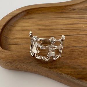 May include: A silver ring featuring a continuous band of stylised figures holding hands in a circle. The metallic band is crafted with cut-out details that define the shapes of the figures, creating an openwork design that wraps around the finger.