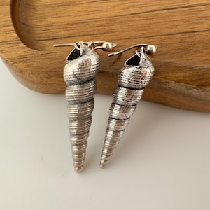 May include: A pair of silver-toned earrings shaped like elongated spiral seashells. Each earring features a detailed, textured surface with horizontal ridges and a hook-style closure at the top, designed for hanging from the earlobe.