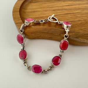 May include: A silver chain bracelet featuring six oval-shaped, faceted red gemstones set in polished silver bezels. The bracelet is secured with a circular toggle clasp and consists of alternating silver links connecting each individual stone setting.