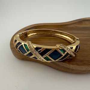May include: Gold-tone hinged bracelet with a geometric design. The bracelet features a pattern of blue, green, and black enamel inlays, accented with clear rhinestones. The bracelet is resting on a wooden surface.
