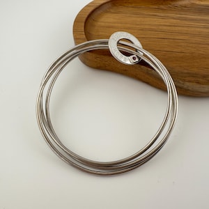 May include: A silver-toned bangle bracelet composed of three interlocking thin circular bands. A small circular charm is attached to the bands, featuring the engraved text "TRUST" and "HOPE" along with a tiny embedded dark red gemstone.