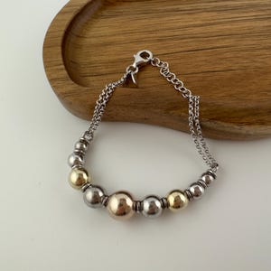 May include: Silver bracelet with a mix of silver and gold-coloured beads. The bracelet has a chain closure and is displayed on a wooden tray. The beads vary in size, with larger gold-coloured beads in the centre.