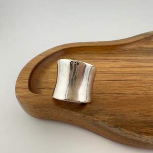 May include: A wide, polished silver ring with a smooth, slightly concave surface and a reflective metallic finish. The band features a curved, architectural shape that tapers slightly towards the centre, showcasing a clean and solid construction.