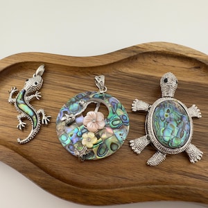 May include: Three decorative pendants on a wooden tray. The pendants include a silver lizard with iridescent accents, a circular pendant with flowers and abalone shell, and a silver turtle with an abalone shell.