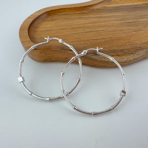 May include: A pair of silver hoop earrings featuring a segmented bamboo-like design with small, smooth cube-shaped accents integrated into the circular frame. The earrings have a polished metallic finish and a hinged closure mechanism at the top.
