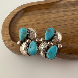 May include: A pair of turquoise and silver earrings. Each earring features three turquoise stones and silver leaf and ball accents. The earrings are set on a wooden tray.