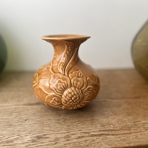 May include: A small, tan-colored ceramic vase with a floral design. The vase has a rounded body and a flared neck. The floral pattern is embossed on the surface, adding texture and detail. The vase is displayed on a wooden surface.