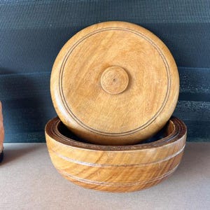 May include: A round, light brown wooden bowl with a matching circular lid. The lid features a small, centered knob and two thin, dark engraved circular lines. The bowl has a smooth, polished finish with visible natural wood grain patterns throughout.