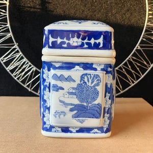 May include: A square ceramic canister with a removable lid, featuring a white base and blue hand-painted landscape scenes. The design includes depictions of trees, mountains, and water, framed by floral patterns on the edges and lid top.