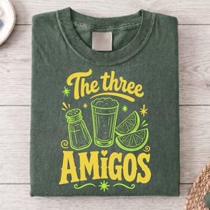 Puede incluir: Camiseta verde oscuro con un diseño gráfico. El diseño presenta el texto "The three AMIGOS" en amarillo, junto con ilustraciones de un salero, una cerveza y rodajas de lima en verde neón.