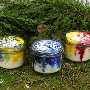 May include: Three glass jar candles with colorful wax. The candles have white pebbles on top and are surrounded by green foliage. The wax colors include yellow, blue, and red, creating a vibrant display. Each candle has a wick.