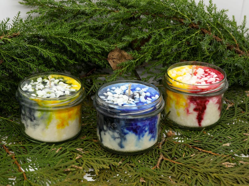 May include: Three glass jar candles with colorful wax. The candles have white pebbles on top and are surrounded by green foliage. The wax colors include yellow, blue, and red, creating a vibrant display. Each candle has a wick.