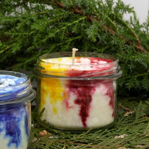 May include: Two glass jar candles with wicks. One candle has a blue and white design, the other has a layered design of yellow, red, and white. The candles are set against a backdrop of green foliage.