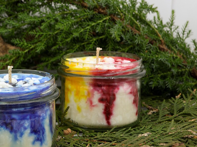 May include: Two glass jar candles with wicks. One candle has a blue and white design, the other has a layered design of yellow, red, and white. The candles are set against a backdrop of green foliage.