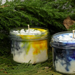 May include: Two glass jar candles with layered wax in white, yellow, and blue hues. Each candle has a wick and is topped with small white stones. The candles are set against a backdrop of green foliage.