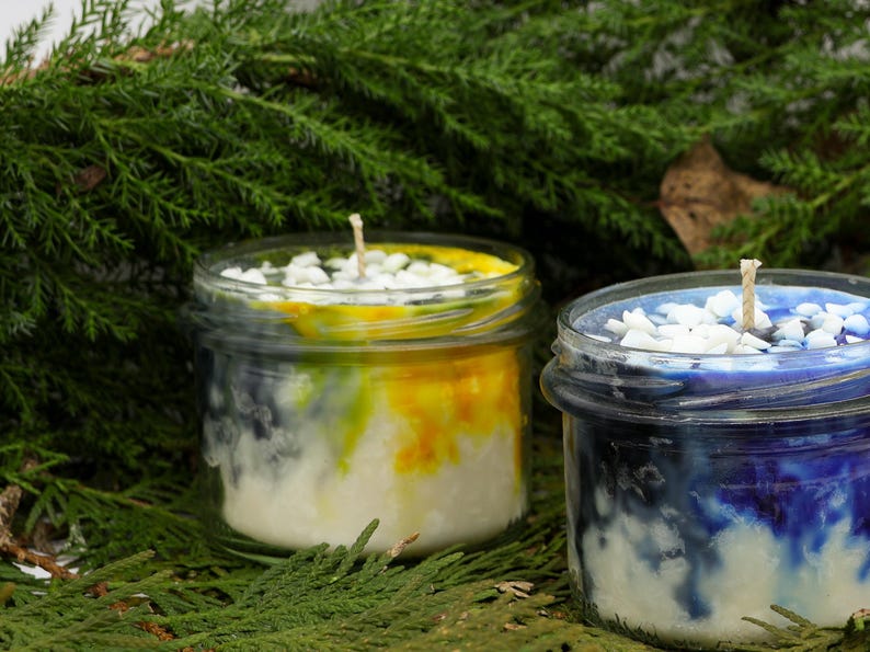 May include: Two glass jar candles with layered wax in white, yellow, and blue hues. Each candle has a wick and is topped with small white stones. The candles are set against a backdrop of green foliage.