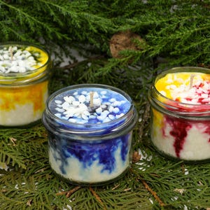 May include: Three glass jar candles with colorful wax and decorative toppings. One candle has yellow and white wax, another has blue and white, and the third has yellow and red wax. The candles sit on a bed of green foliage.