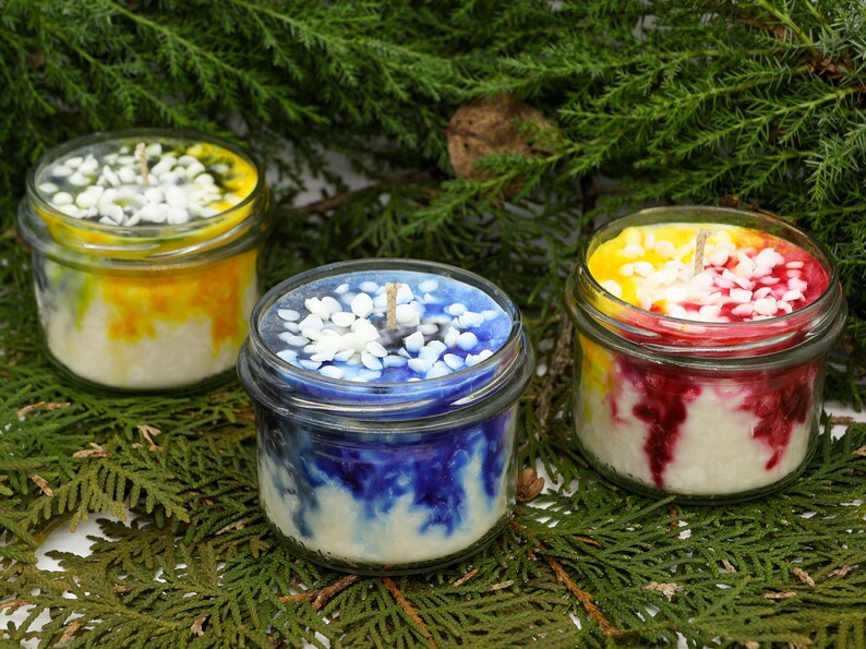 May include: Three glass jar candles with colorful wax and decorative toppings. One candle has yellow and white wax, another has blue and white, and the third has yellow and red wax. The candles sit on a bed of green foliage.