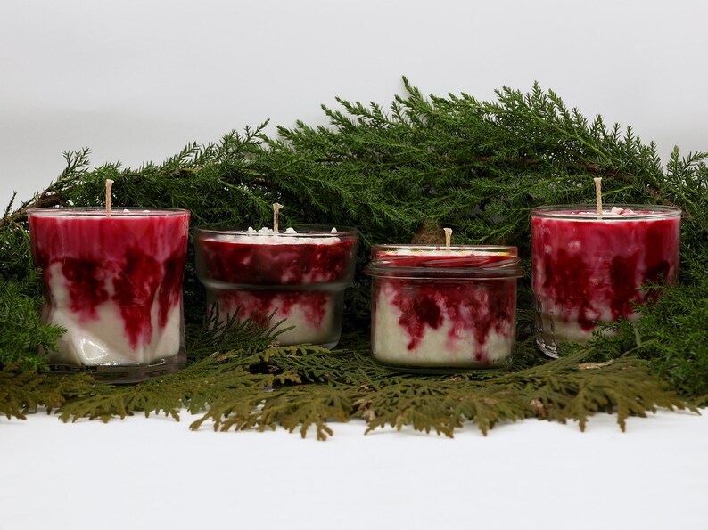 May include: Four candles with red and white marbled wax are displayed against a backdrop of green foliage. The candles are in clear glass containers, with one in a jar with a gold lid. Each candle has a single wick.