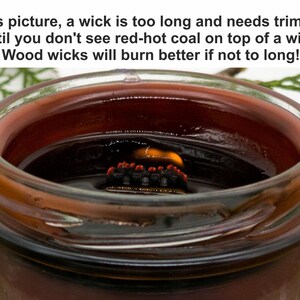 May include: A close-up of a candle in a clear glass jar. The candle has a long, charred wooden wick with red-hot coal on top. The candle wax is a dark reddish-brown color. The text in the image says the wick is too long and needs trimming.