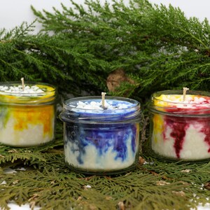 May include: Three small, glass jar candles with colorful wax. One candle has yellow and orange, another has blue and white, and the third has red and yellow. Each candle has a white wick and is surrounded by greenery.