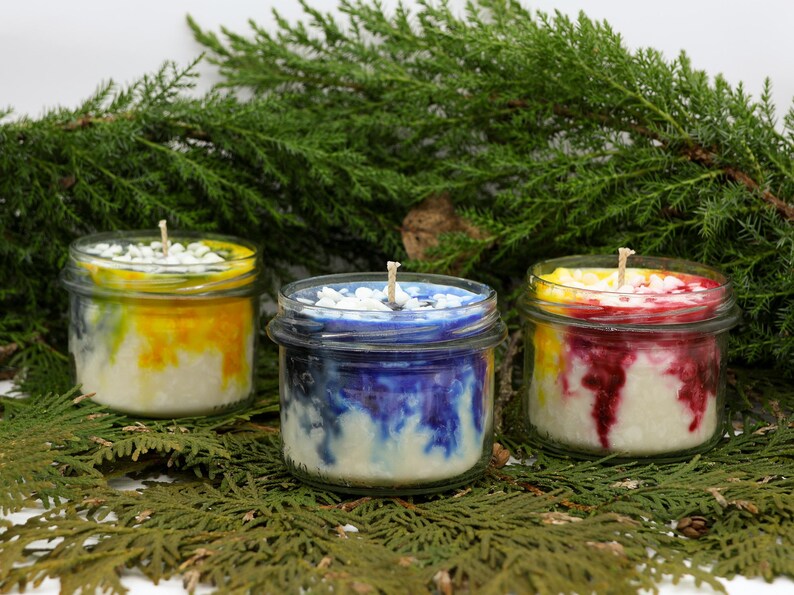 May include: Three small, glass jar candles with colorful wax. One candle has yellow and orange, another has blue and white, and the third has red and yellow. Each candle has a white wick and is surrounded by greenery.