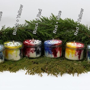 May include: Six glass jar candles with colorful wax in shades of blue, yellow, and red, with black accents. The candles are arranged in front of a backdrop of green foliage. The labels on the candles read "Black + Blue", "Black + Yellow", "Black + Red", "Red + Blue", "Red + Yellow", and "Blue + Yellow".
