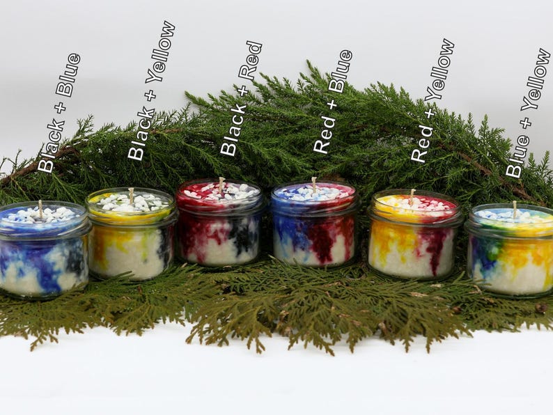 May include: Six glass jar candles with colorful wax in shades of blue, yellow, and red, with black accents. The candles are arranged in front of a backdrop of green foliage. The labels on the candles read "Black + Blue", "Black + Yellow", "Black + Red", "Red + Blue", "Red + Yellow", and "Blue + Yellow".