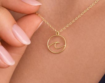 May include: A gold-colored necklace featuring a circular pendant with a wave design. The wave is a simple, elegant line drawing within the circle. The necklace is on a delicate chain.