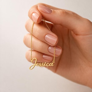 Può includere: Una collana color oro con il nome "Jessica" in corsivo. La collana è tenuta da una mano con unghie curate. La catena è delicata e il ciondolo è una singola parola.