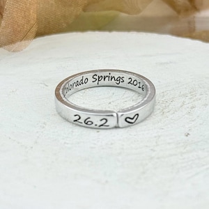 Puede incluir: Un anillo plateado con la inscripción grabada "Colorado Springs 2016" y "26.2" con un símbolo de corazón. El anillo se muestra sobre una superficie blanca y texturizada.