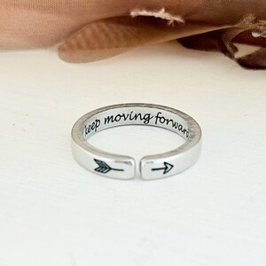 May include: A silver-toned open band ring featuring the engraved text "keep moving forward" along the inner surface, with a small arrow symbol engraved on the outer band near the opening, creating a simple and functional piece of jewelry.