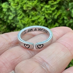 May include: A silver ring with the words "I am a Writer" engraved on the band. The ring has two small heart cutouts on either side of the band.