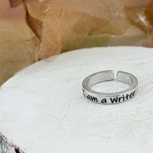 May include: Silver adjustable ring with the inscription "I am a Writer". The ring is displayed on a white, textured, wooden surface. The background features soft, blurred colors.