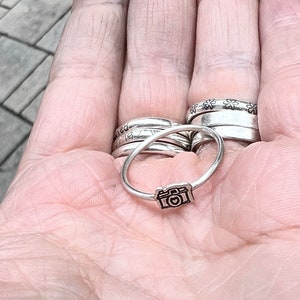 Może przedstawiać: Srebrny pierścionek z małym wisiorkiem w kształcie aparatu fotograficznego. Pierścionek jest noszony na palcu, wraz z kilkoma innymi srebrnymi pierścionkami.