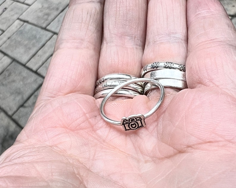 Może przedstawiać: Srebrny pierścionek z małym wisiorkiem w kształcie aparatu fotograficznego. Pierścionek jest noszony na palcu, wraz z kilkoma innymi srebrnymi pierścionkami.