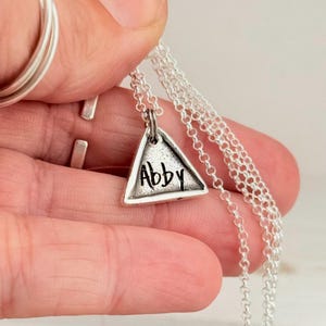 May include: A silver necklace featuring a triangular pendant with the name "Abby" inscribed in a handwritten style. The pendant hangs from a delicate chain. The necklace is held in a hand, with other silver rings visible in the background.
