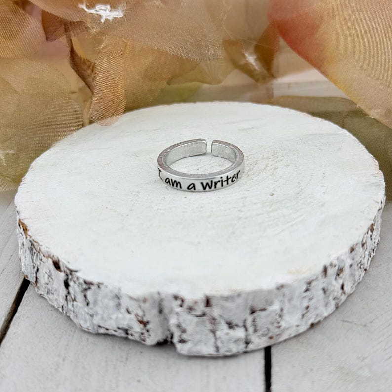 May include: Silver adjustable ring with the inscription "I am a Writer". The ring is displayed on a white, textured, wooden surface. The background features soft, blurred colors.
