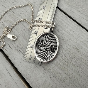 May include: Silver fingerprint pendant necklace with a detailed fingerprint design. The pendant is oval-shaped and hangs from a delicate silver chain. A ruler is placed next to the pendant, showing measurements in millimeters.