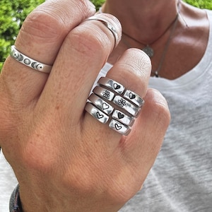 K&ouml;nnte beinhalten: Ein Stapel von f&uuml;nf silbernen Ringen mit verschiedenen eingravierten Symbolen. Die Symbole beinhalten Herzen, Blumen und Sterne.