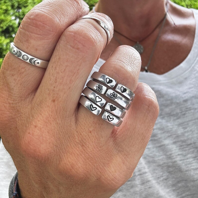 K&ouml;nnte beinhalten: Ein Stapel von f&uuml;nf silbernen Ringen mit verschiedenen eingravierten Symbolen. Die Symbole beinhalten Herzen, Blumen und Sterne.