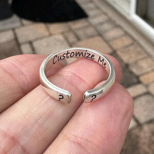 K&ouml;nnte beinhalten: Ein silberner Ring mit der Gravur "Customize Me" auf der Band. Der Ring ist an einer Seite offen und hat an jedem Ende ein Fragezeichen.
