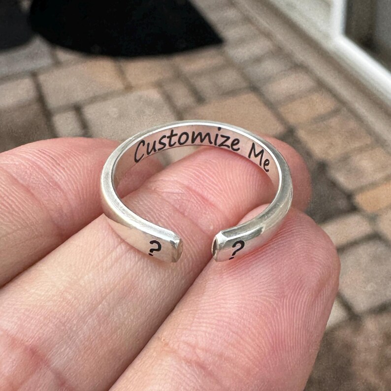 K&ouml;nnte beinhalten: Ein silberner Ring mit der Gravur "Customize Me" auf der Band. Der Ring ist an einer Seite offen und hat an jedem Ende ein Fragezeichen.